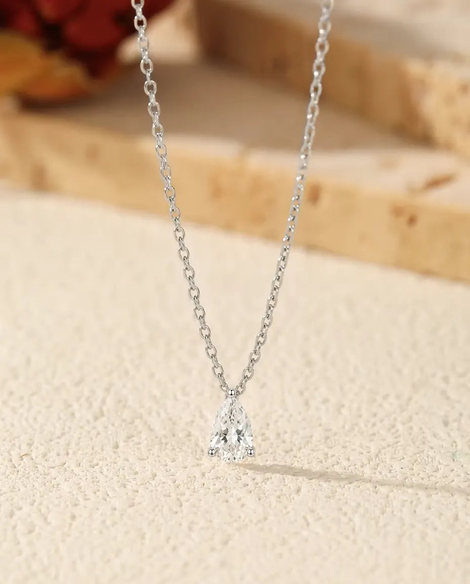 1 ct Pear Moissanite Necklace in 925 Sterling Silver - pendant resting on a cream surface beside a travertine slab
