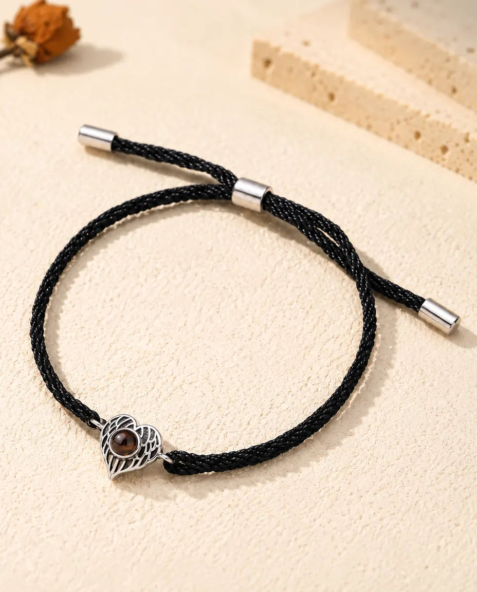Personalised photo projection bracelet with a silver tone angel wing heart on a black rope cord, lying on cream plaster