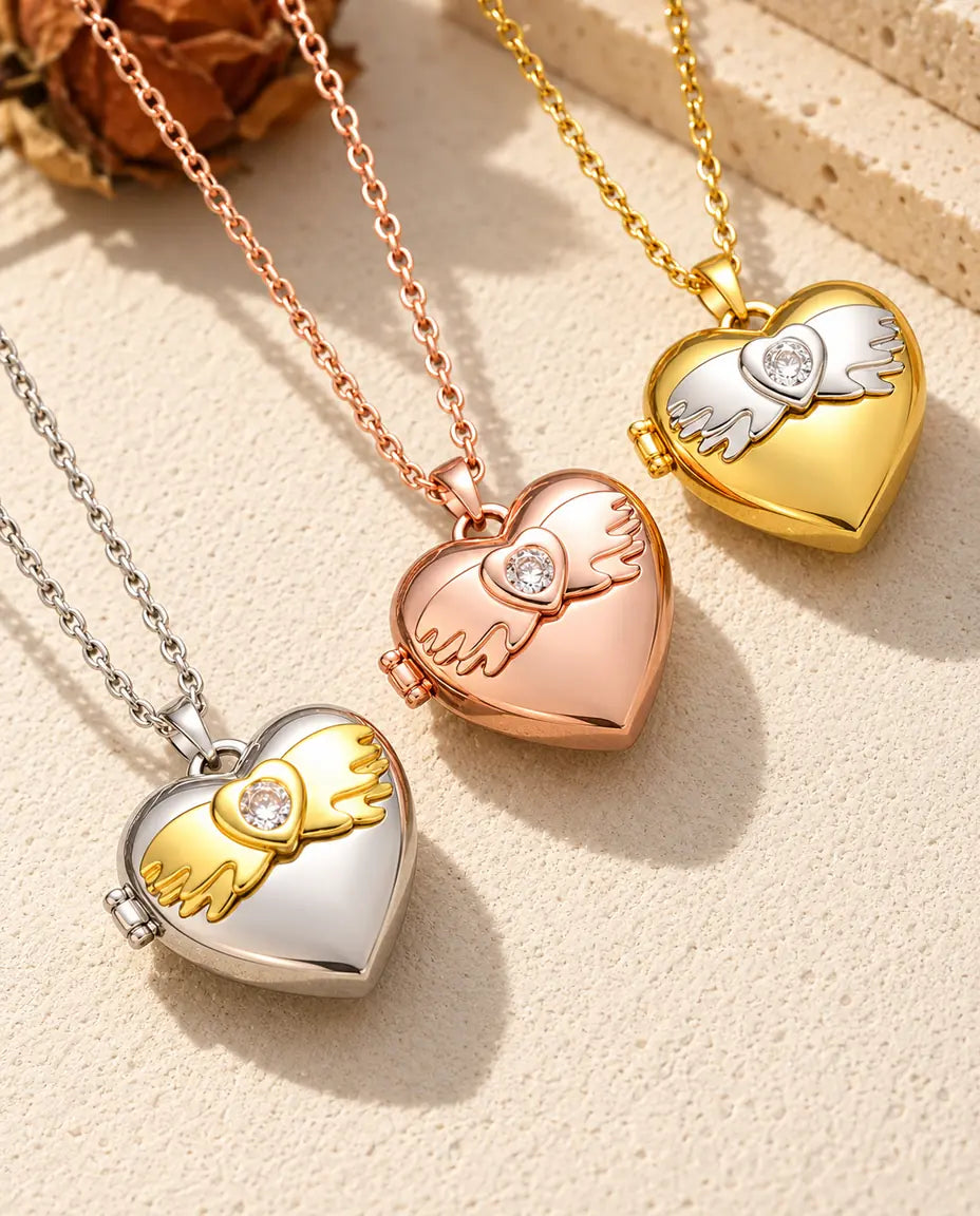 Angel wings heart photo lockets in silver tone, rose gold tone and gold tone laid diagonally on cream plaster