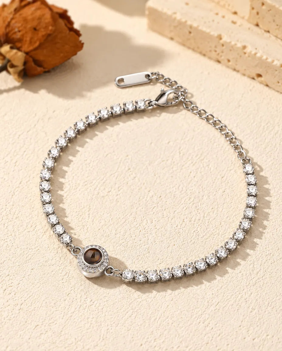 Personalised photo projection tennis bracelet in silver tone with a halo charm, lying on cream plaster