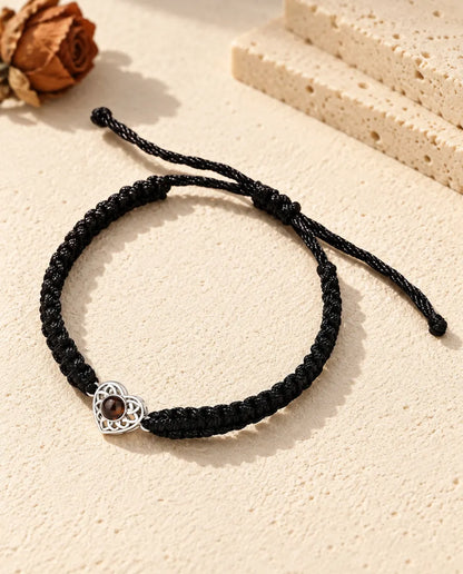 Personalised photo projection bracelet with a silver tone openwork heart charm on a black braided cord, lying on cream plaster