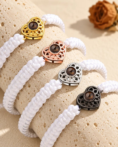 Four white braided cord bracelets with gold tone, rose gold tone, silver tone and black heart charms on a travertine block
