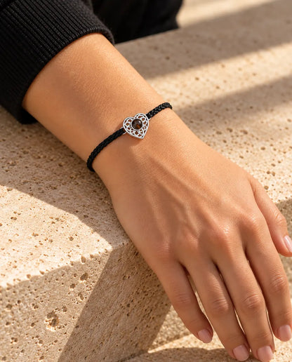 Woman wearing the black thin braided cord bracelet with a silver tone heart charm on her wrist in warm sunlight