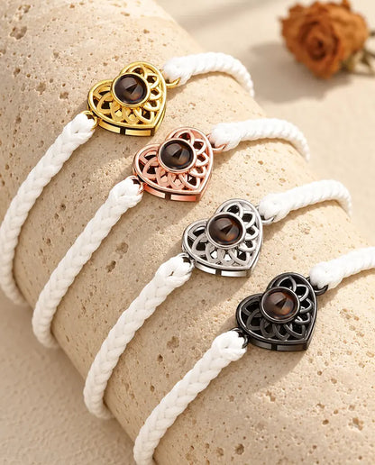 Four white thin braided cord bracelets with gold tone, rose gold tone, silver tone and black heart charms on a travertine block
