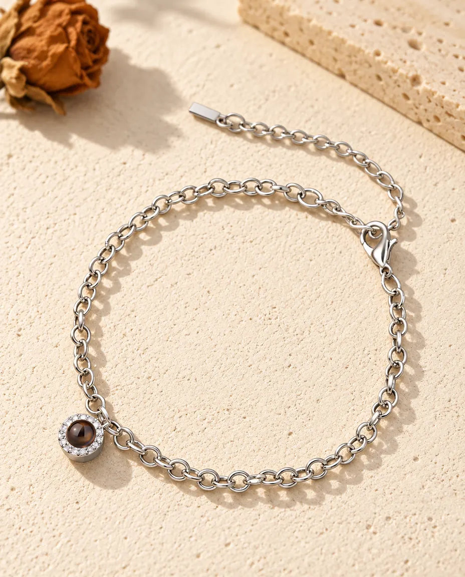 Personalised photo projection bracelet in silver tone with a tiny pave circle charm on a fine chain, lying on cream plaster