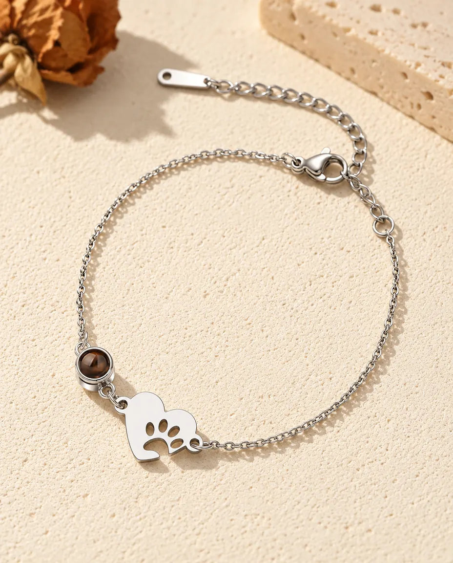 Personalised photo projection bracelet with a silver tone paw print heart tag and projection stone on a fine chain, lying on cream plaster