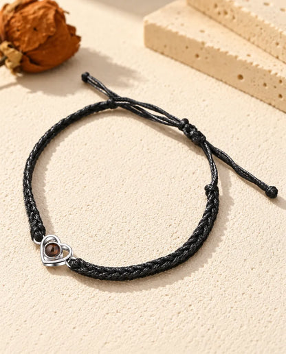 Personalised 925 sterling silver open heart photo projection bracelet on a black braided cord, lying on cream plaster